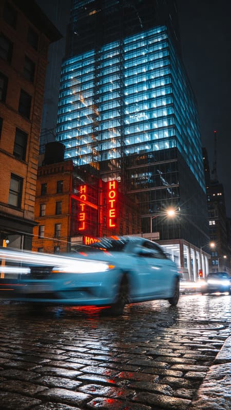 Double exposure photograph of a bustling city street at night. The foreground features a wet cobblestone street with a light blue car in motion, leaving light trails. The background is a complex overlay of a modern glass skyscraper and an older brick building, creating an abstract, layered effect. Red neon "HOTEL" signs are visible through the layers. The lighting is dark and moody, with reflections and light trails from streetlights and car headlights. The colors are dominated by cool blues from the skyscraper and warm oranges/browns from the brick building and neon signs. High contrast, urban atmosphere, with a grimy, gritty texture. Photorealistic, editorial photography.