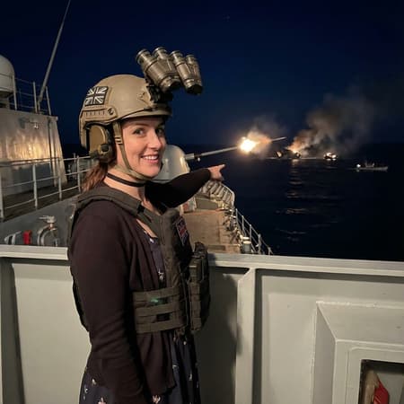 she's wearing by herself in the same outfit, but instead she wears a modern ballistic helmet with a british flag patch on the side equipped with quanods GPNVGS that are flipped up, and a plate carrier over her cardigan and dress, and choker, she's standing atop a warship, pointing out at see, directing the battleship guns as they fire on small civilian boats in the distance, there is wreckage from destroyed ones burning with smoke billowing from them, she look back with a smile, at night