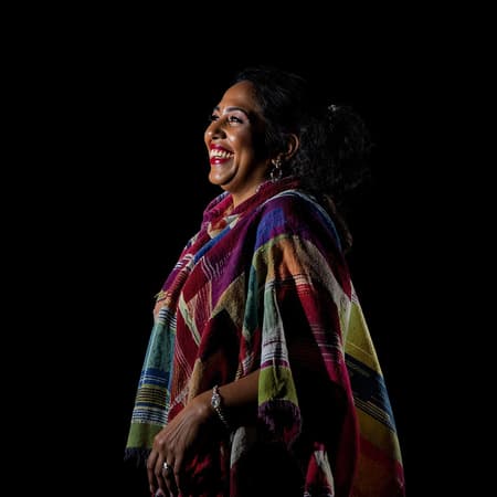 Dark background dramatic lighting with backlight and rim lighting features a 44-year-old Mexican woman captured from the side, her full body standing gracefully. She's laughing joyously, resembling a unique beauty, draped in a colorful, translucent poncho that plays with the light. Silver jewelry adorns her, adding elegance. The image is of the highest quality, shot in 4k, and 8k ultra-high resolution, with raw HDR capturing every detail. Her skin reveals intricate textures and natural imperfections, her eyes detailed and glossy, highlighting her expressive gaze. The scene is devoid of unnecessary elements like text or extra fingers, focusing purely on the natural beauty and authenticity of the subject.