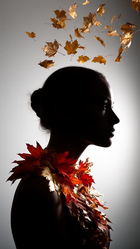 Woman's Silhouette Adorned With Autumn Leaves Photorealistic studio shot of a woman's silhouette in profile, facing right. Her head and upper body are visible, rendered as a stark black shadow against a light grey gradient background. Autumn leaves in vibrant shades of red, orange, brown, and metallic gold are artfully arranged on her neck and shoulder, creating a natural garland. Additional autumn leaves, including some golden ones, are scattered and falling above and around her head, appearing to float in the air. The lighting is dramatic and high-contrast, with a strong light source from the right illuminating the leaves and creating a subtle glow on the background. The mood is ethereal and artistic, celebrating the beauty of autumn.