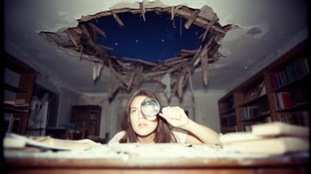 70s vintage film photography, a dreamlike and cosmic scene in a ruined library. A young woman with a sharp, intellectual face is lying on a table, directly beneath a hole where the ceiling has collapsed. Through the hole, a patch of night sky is visible, but out of focus. She holds a broken magnifying glass over one eye, not looking up, but pointed blankly at the splintered rafters. The heavy flash overexposes the lens and the side of her face, creating a stark, glassy highlight. The palette is a faded, starlit indigo and plaster-dust white. The blur of fallen books looks like distant nebulae. She is studying the intimate, ruined architecture of her own collapsed universe.