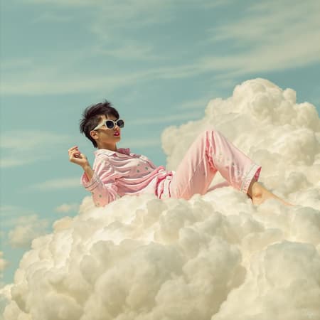 An imaginative photo of an eccentric woman lounging atop billowing white clouds, wearing oversized sunglasses and pastel pink pajamas adorned with green polka dots; her short dark hair styled in a playful pixie cut. The backdrop is a serene baby blue sky with whimsical cloud formations, evoking a dreamlike ambiance. Captured in the style of Jimmy Marble, the image features muted earthy colors and soft tones, adding a whimsical yet grounded charm.