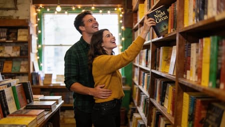 Happy Couple Laughing In Cozy Bookstore A happy couple in an old bookstore, a woman in a mustard yellow sweater and black jeans reaching for a book on a high shelf, with a man in a dark green plaid shirt and dark pants standing behind her with his arm around her waist, both looking up and laughing. Photorealistic, warm, natural light from a front window and string lights, cozy and joyful atmosphere, soft focus background. Rich browns, greens, and yellows, with diverse book spine colors. "The Night Circus" visible on the book the woman is holding.