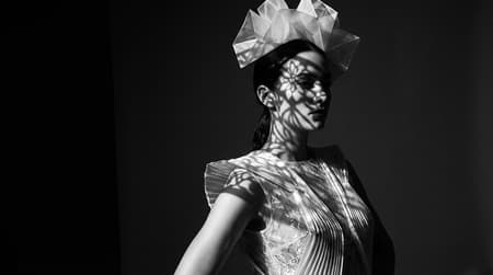 A high-fashion fine art portrait of a female model posed elegantly in three-quarter profile, sculptural and poised. The model wears an avant-garde, structured dress with delicate, almost lace, fabric textures and a dramatic, architectural headpiece composed of pleated, faceted forms surrounding the head.

Complex projected light patterns from an unseen external source cast intricate organic and floral shadows across the model’s face, body, dress, and headwear. The projections interact with the folds, seams, and contours of the garments, revealing fine material detail and depth. Light and shadow wrap around the figure, creating a strong chiaroscuro effect.

Black-and-white cinematic photography with deep blacks, luminous highlights, and rich midtones. Extreme clarity and micro-detail in fabric weave, pleats, and surface texture. Sharp focus throughout the subject, no softness, high contrast without crushing shadows.

Minimal dark studio background, subtle atmospheric falloff. The mood is sculptural, timeless, and editorial — inspired by classic fashion photography and contemporary light-art installations.

chiaroscuro lighting, projection mapping, high-contrast monochrome, fine art fashion photography, sculptural portrait, ultra-sharp detail, studio control, dramatic shadows, medium format look, editorial precision

