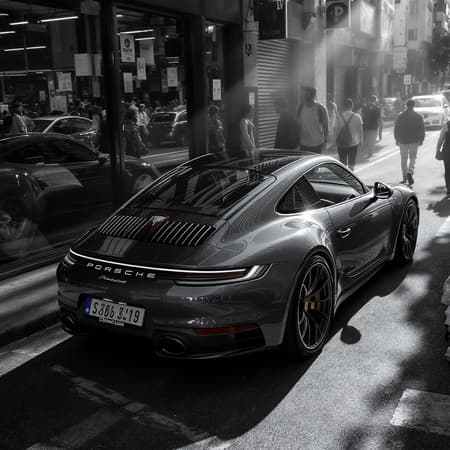 Porsche 911 :: Intense, directional light casting long shadows :: Vibrant background filled with bustling activity :: Car prominently displayed in the frame's center :: Lively urban street teeming with pedestrians and vehicles :: Car's reflection glimmering in a storefront window :: Detailed close-up of exhaust and diffuser :: People walking in front, emphasizing car's presence :: Black and white filter for a gritty, dynamic feel :: Elevated angle capturing the street's liveliness :: Prime lens ensuring sharpness and smooth bokeh :: Ultra detailed and photorealistic --ar 3:2 --s 50.