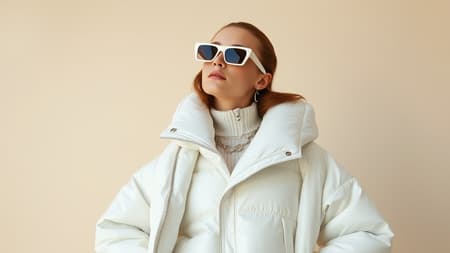 The image captures a woman posed against a soft beige background, epitomizing a Tumblr aesthetic. She is clad in a striking off-white ensemble featuring a glossy reflective puffer jacket layered with a chic poncho. This look echoes a popular online trend and has gained traction on the design-forward website Dezeen, making waves in their 2024 fashion showcase. Her accessories include a trendy mac, chic 8.0 LV sunglasses, and a sculptural piece designed by Mike and Wojtek Fus. The ensemble is a testament to avant-garde style, poised to captivate attention and embody the forefront of contemporary fashion.