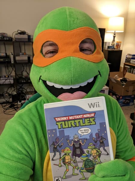 Photorealistic close-up of a person in a bright green Teenage Mutant Ninja Turtles costume with an orange mask, holding up a custom-designed Wii game case titled "SKINNY MUTANT NINJA TURTLES". The person's eyes are visible through the mask's eyeholes, and a wide, happy smile from the costume mouth reveals white teeth and a pink tongue. The game case cover features cartoonish, skinny versions of the four turtles, Shredder, and Foot Clan ninjas, with a speech bubble saying "You skinny reptiles!". The background is a blurred, messy room with console shelves and electronics. Indoor lighting, vibrant colors, playful and humorous mood.