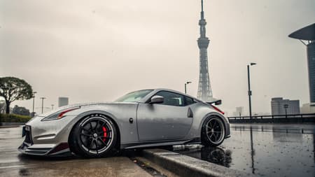 Sideview of a Nissan 370z nismo in silver and extremly lowered with an airlift system and deep rims near the tokyo sky tree on a rainy day